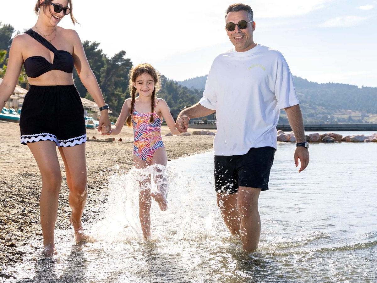 Two adults and a child walking hand in hand through ankle-deep water at the shoreline, with hills across the bay.