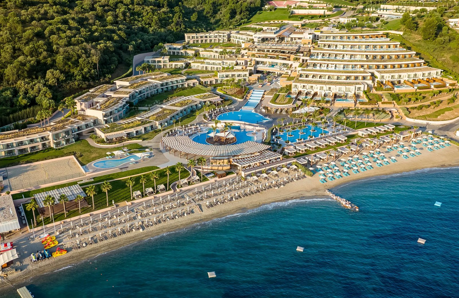 Miraggio Thermal Spa Resort stepped down a wooded hillside in curved white terraces to its pools and long sandy beach, with the Aegean in the foreground.