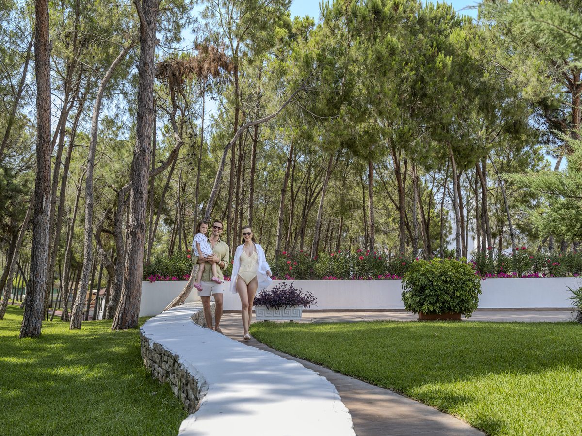 Paved path curving between mown lawns and planted beds under tall pine trees, with white resort buildings beyond.