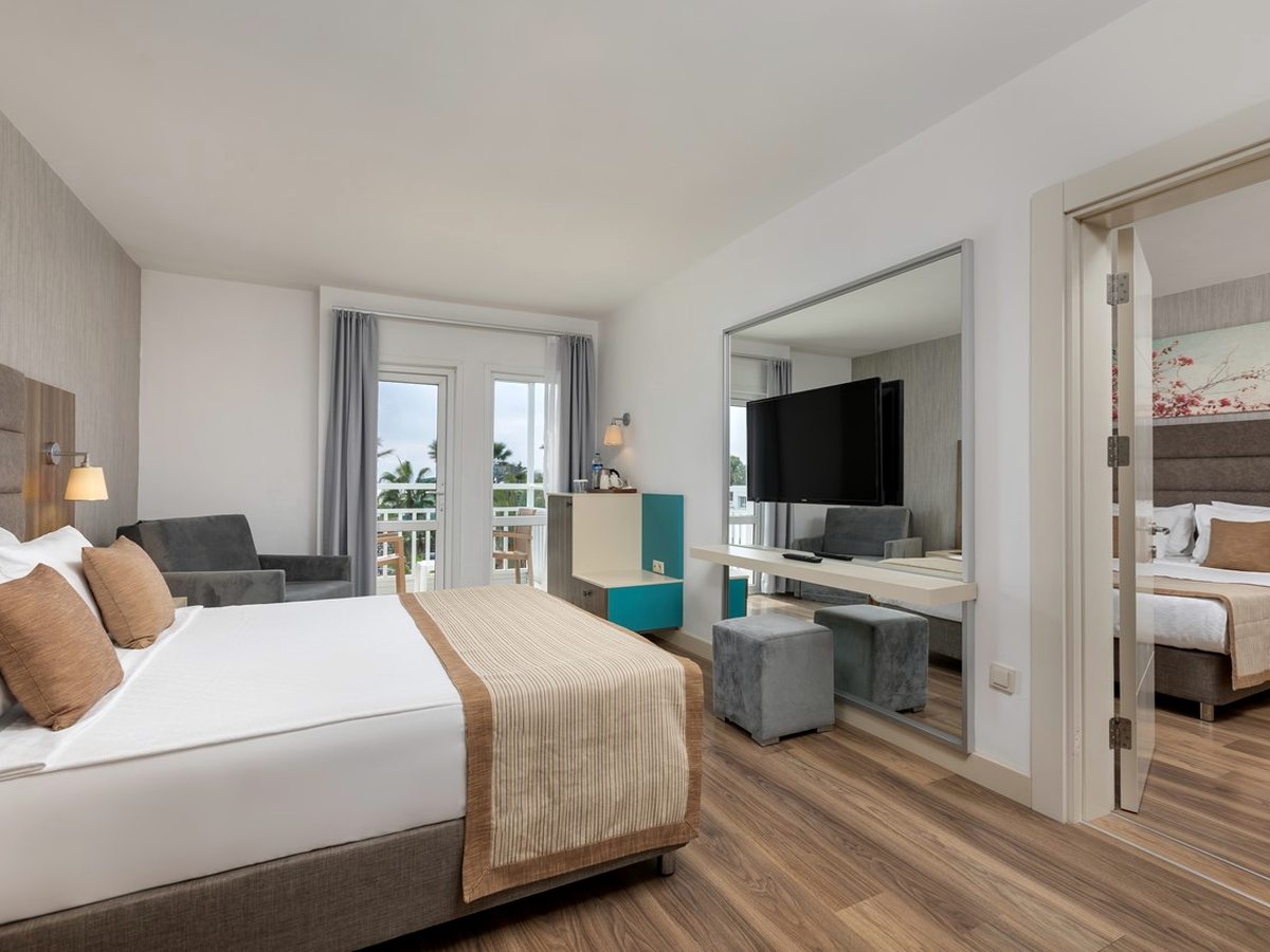 Room with a double bed, grey armchair and balcony doors, with a second bedroom through an open door to one side.
