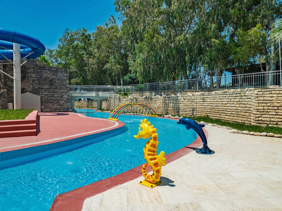Shallow winding children's channel with a yellow seahorse sprayer, a dolphin sprayer and a rainbow arch of water jets, with a slide behind.