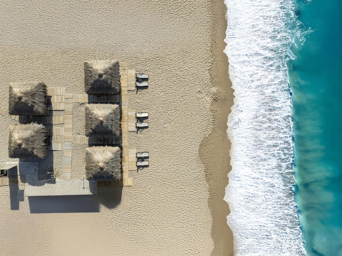 Aerial view of thatched cabanas and sun loungers set out in rows on pale sand, with turquoise shallows breaking on the shore.