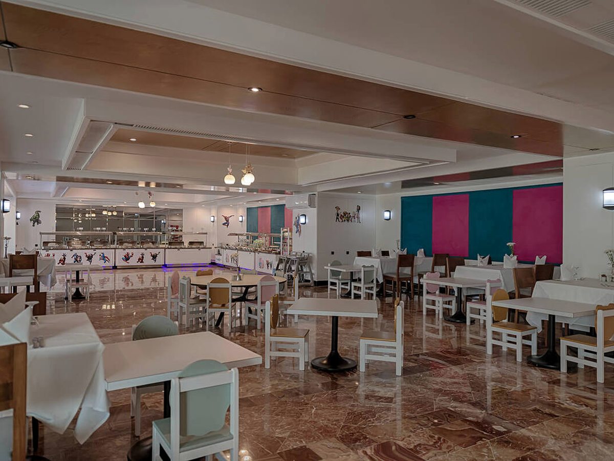 Children's dining room with low wooden chairs at white tables, cartoon-character panels along the servery and teal and pink wall panels.