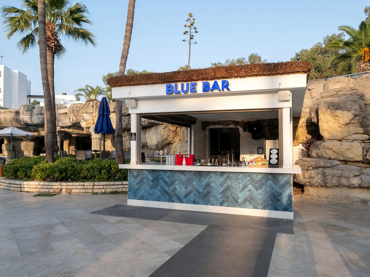 Poolside bar kiosk with a service counter under a BLUE BAR sign, set among palms and artificial rockwork.