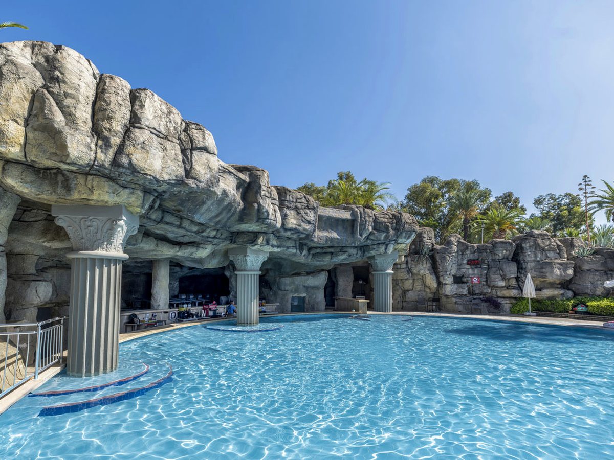 Outdoor pool set against artificial rockwork with white columns, palms and sun loungers around the edge.