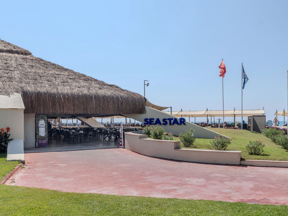 Thatched open-sided beachside restaurant with dark tables and chairs under the roof, a SEASTAR sign on the ramp wall and flags on the lawn outside.
