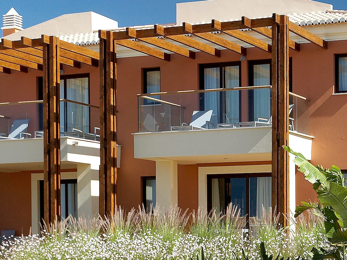 A terracotta apartment block with glass-fronted balconies under heavy wooden pergolas, tall grasses flowering in front.