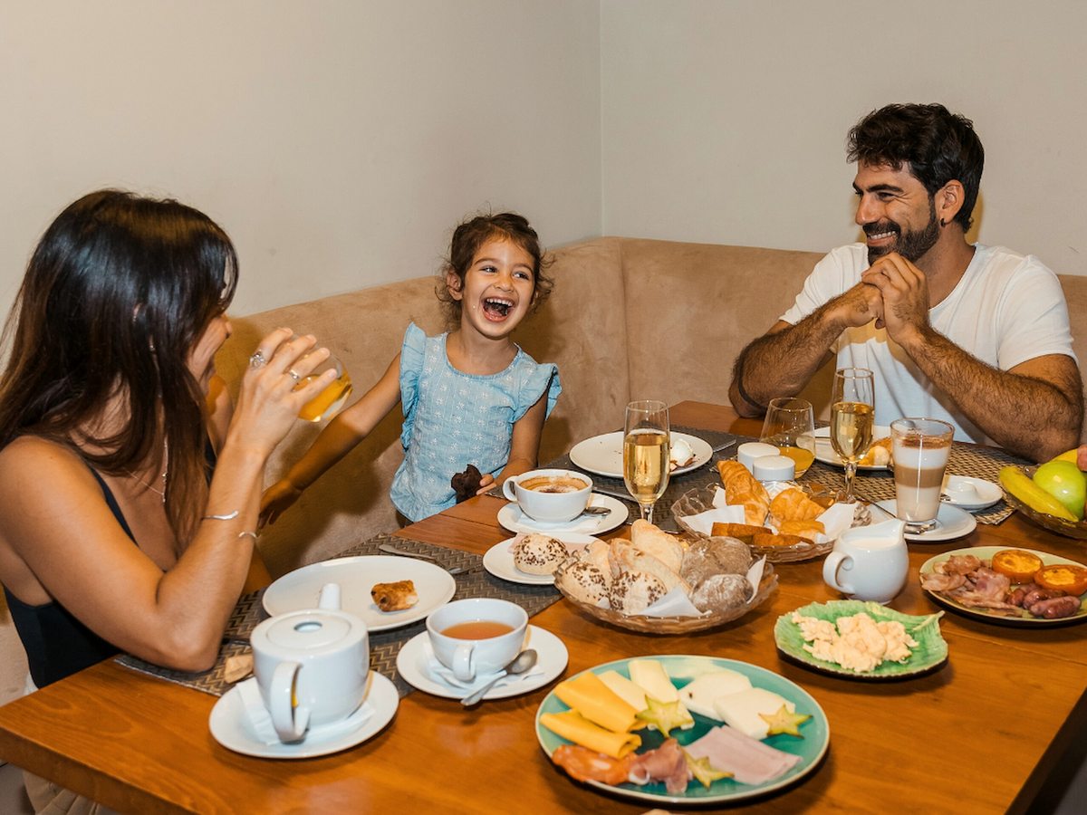 A couple and a young girl laughing around a wooden table spread with pastries, bread, cheese, ham, coffee and juice.