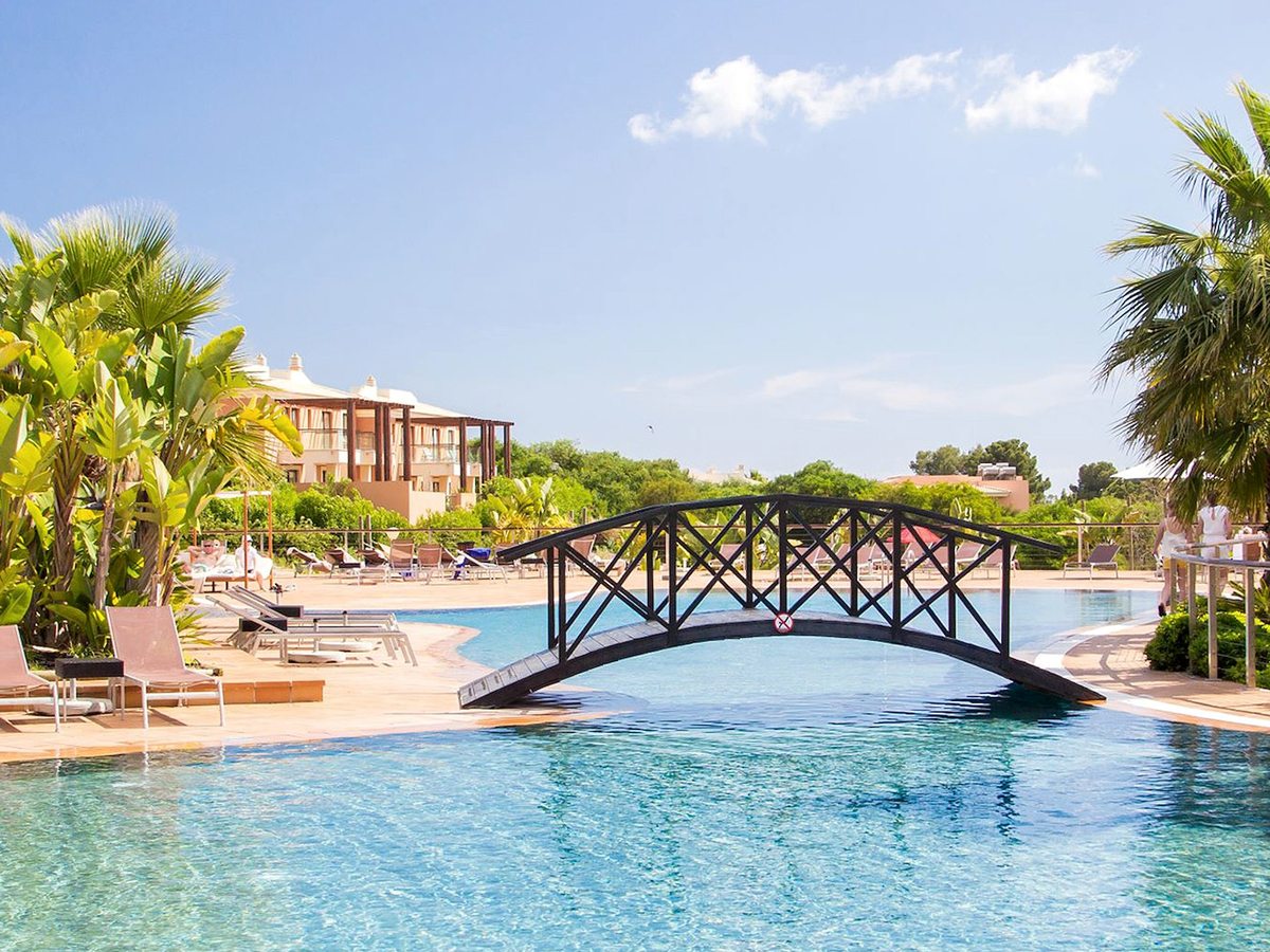 A black-railed wooden bridge arching over the blue pool, with sun loungers, palms and resort buildings behind.