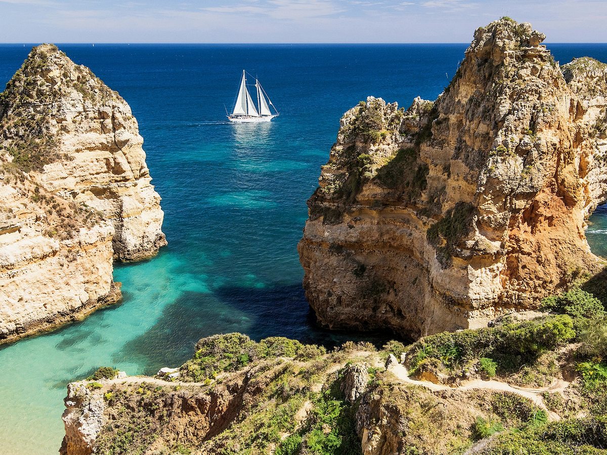 Golden rock stacks rising from clear turquoise and deep blue sea, with a sailing boat passing between them and a path along the cliff top.