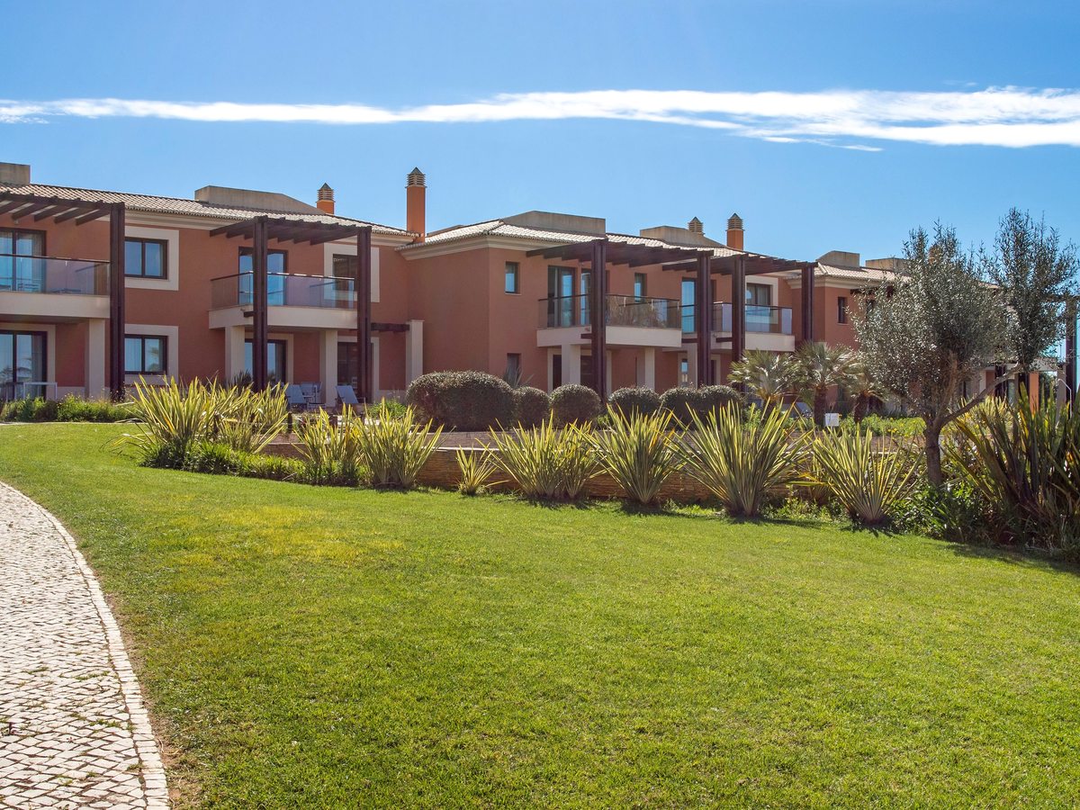 A wide lawn edged with spiky planting in front of a row of two-storey terracotta apartment buildings with wooden pergolas, under a blue sky.