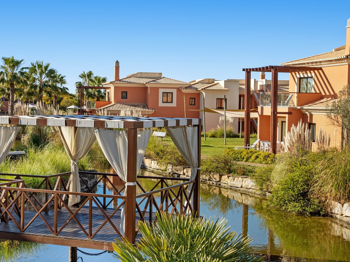 A wooden deck with a curtained pergola built out over a narrow garden waterway, with terracotta resort buildings and palms behind.