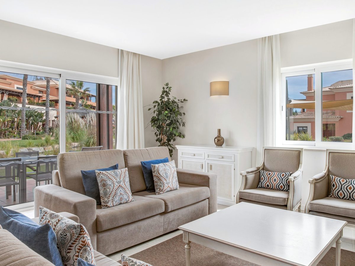 A living room with beige sofas, patterned cushions, a white sideboard and two armchairs, with sliding doors onto a terrace and garden.