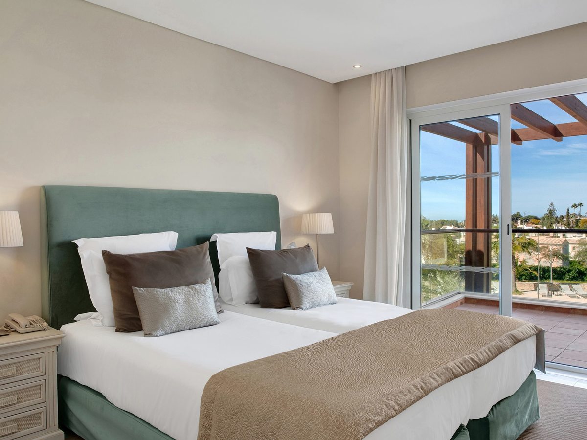 A bedroom with twin beds pushed together under a green upholstered headboard, bedside lamps and glass doors onto a pergola-shaded balcony.