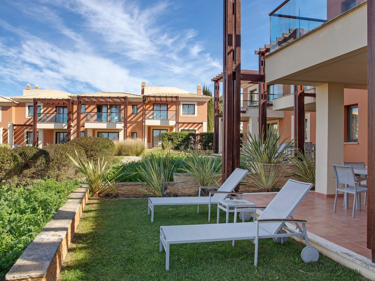 Sun loungers on a lawn beside a ground-floor terrace with a dining table, with terracotta apartment buildings and planted beds behind.