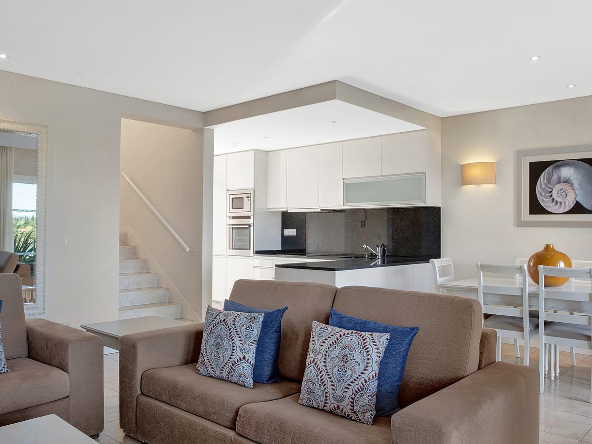 An open-plan living room with two beige sofas, a white dining table, a white and black kitchen behind and a staircase leading up.