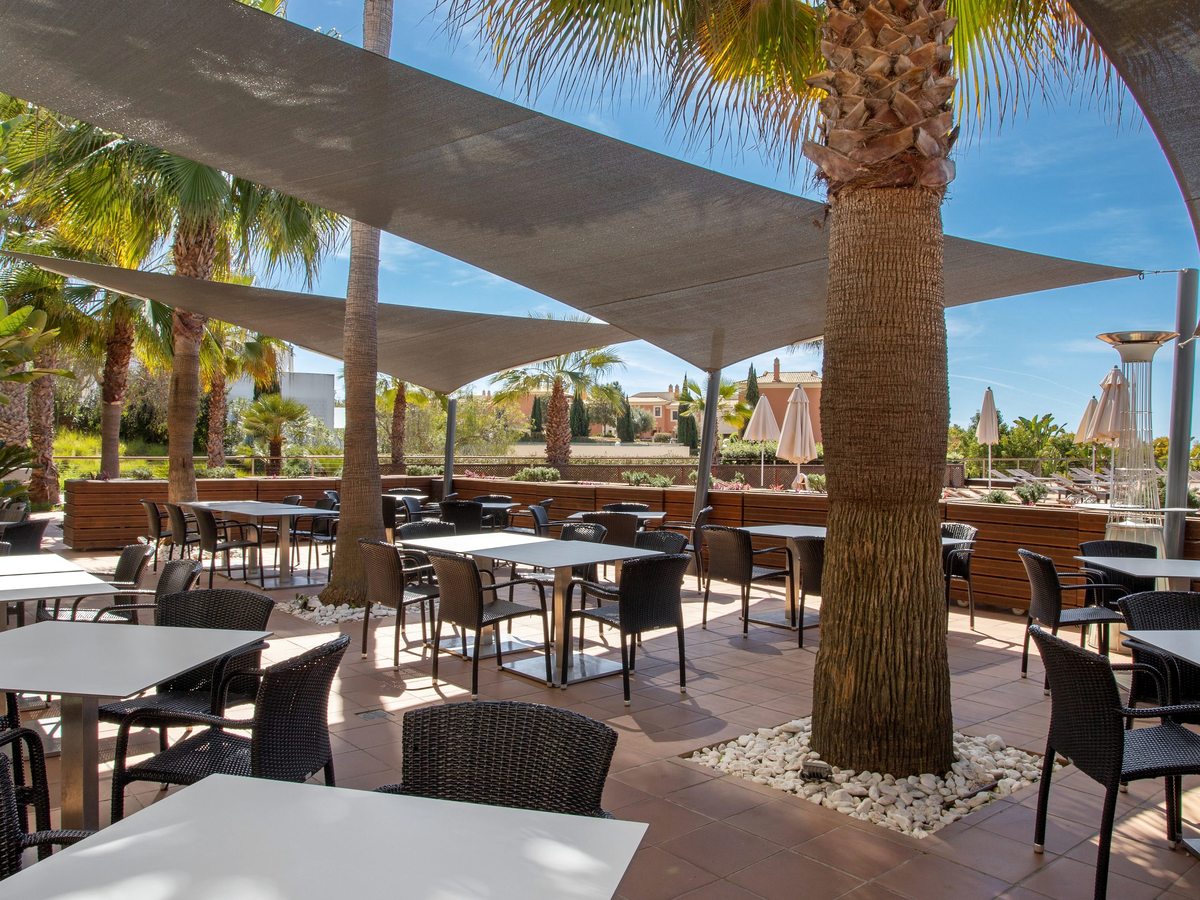 An outdoor restaurant terrace with white tables and dark wicker chairs under grey shade sails, palms growing up through the paving.