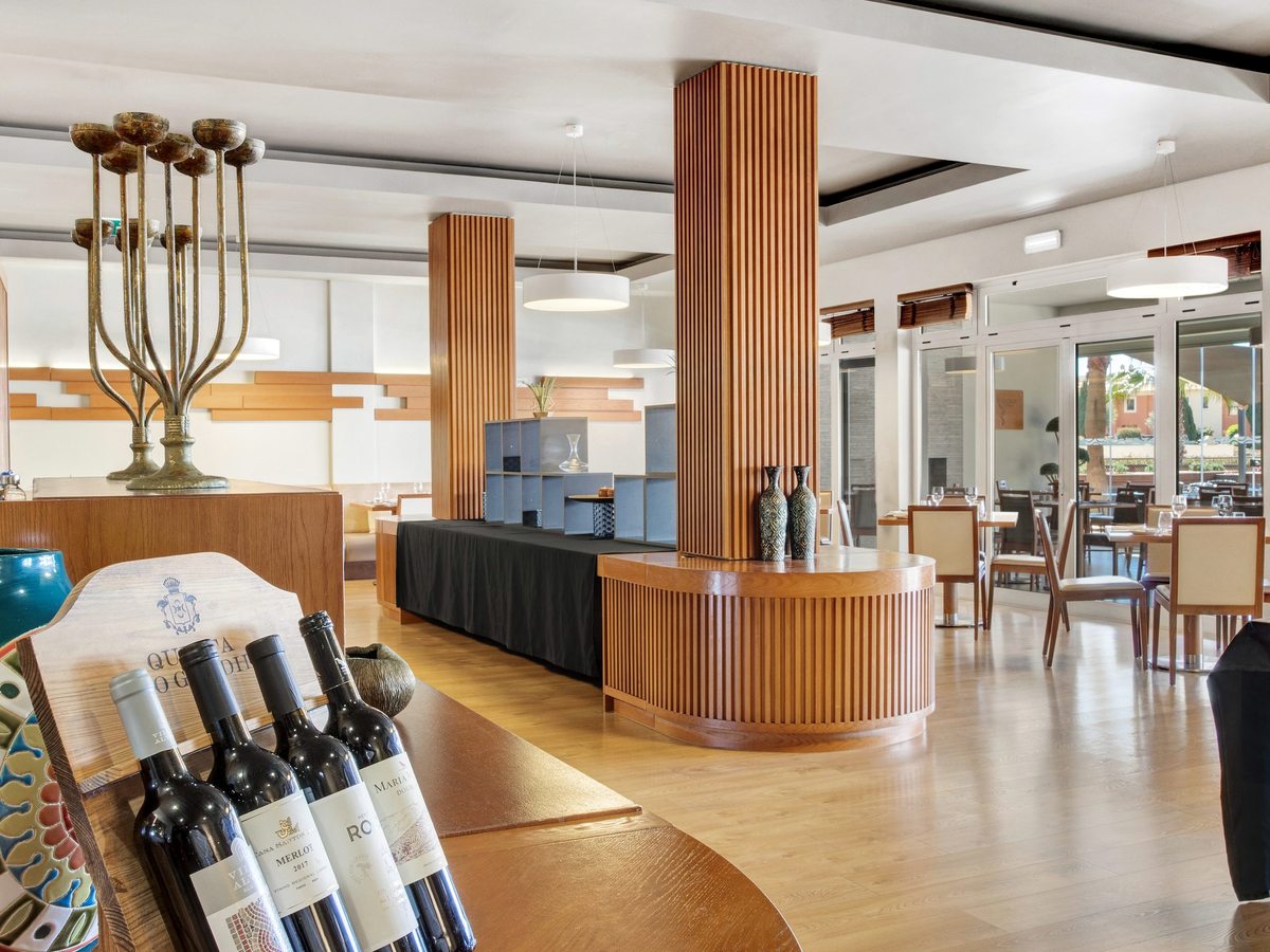 A restaurant dining room with pale walls, wooden slatted pillars, a curved wooden serving counter and set tables beside glass doors to a terrace.