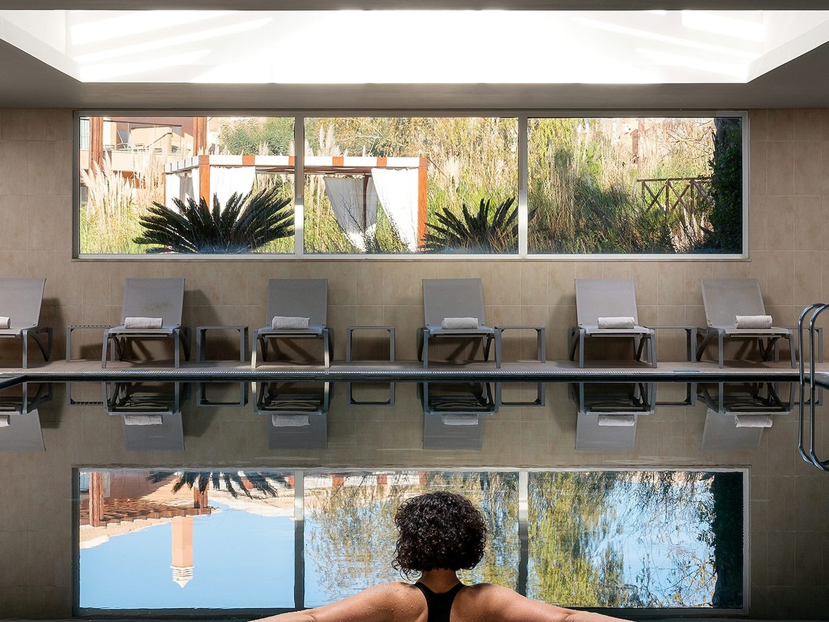 An indoor pool in a tiled room with a row of grey loungers along one side and a long window onto the garden.