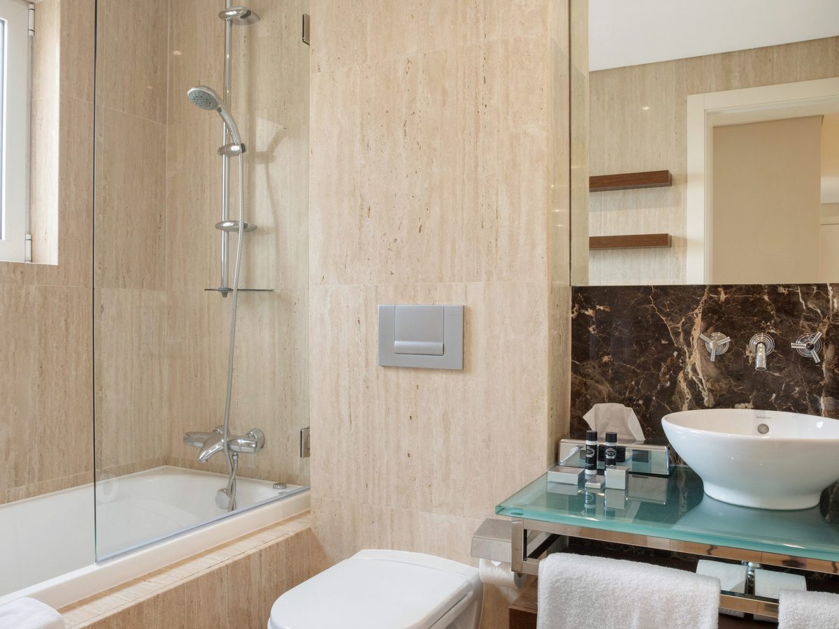 A travertine-tiled bathroom with a bath and shower behind a glass screen, a WC and a round basin on a glass shelf with a marble splashback.