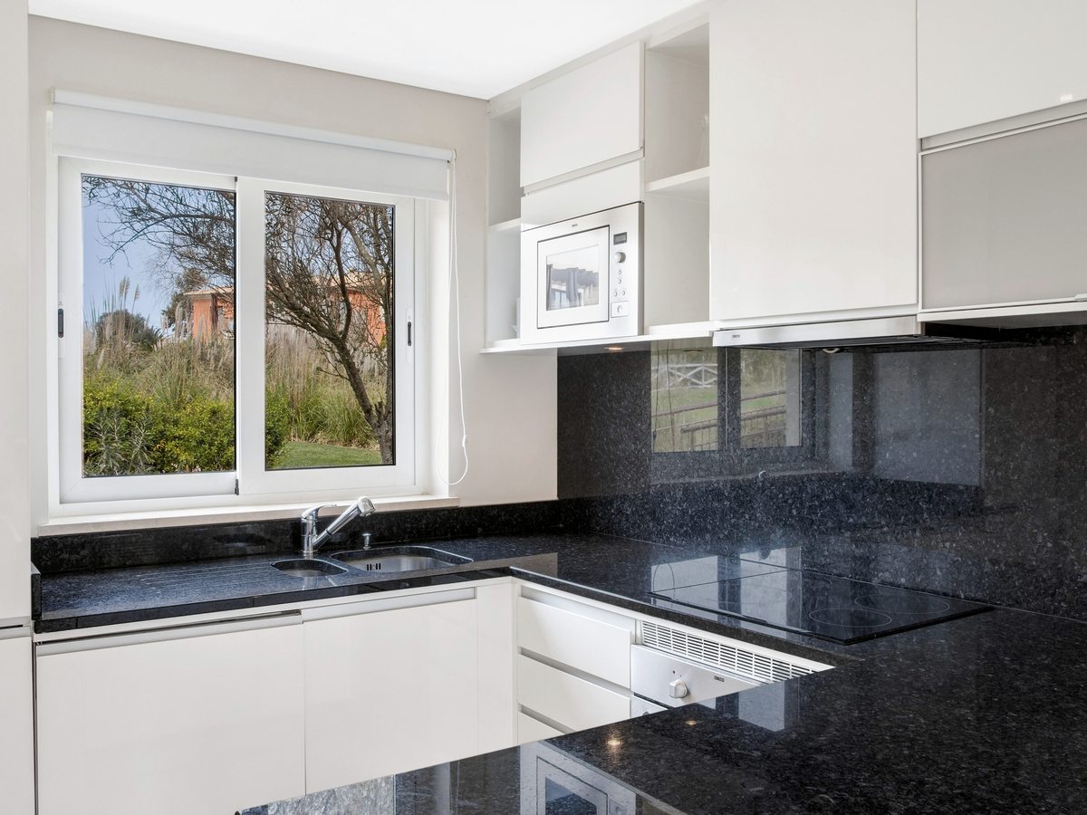 A white fitted kitchen with black granite worktops, a hob and extractor, built-in microwave and oven and a window onto the garden.