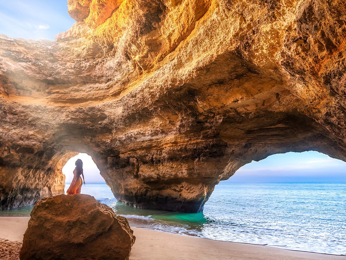The inside of a sandstone sea cave with two arches opening onto a sandy beach and calm sea, a figure standing on a rock.