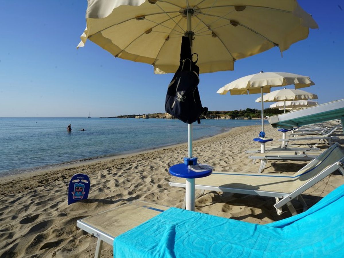 Sun loungers under cream parasols on pale sand at the water's edge, with calm blue sea and a low rocky headland in the distance.