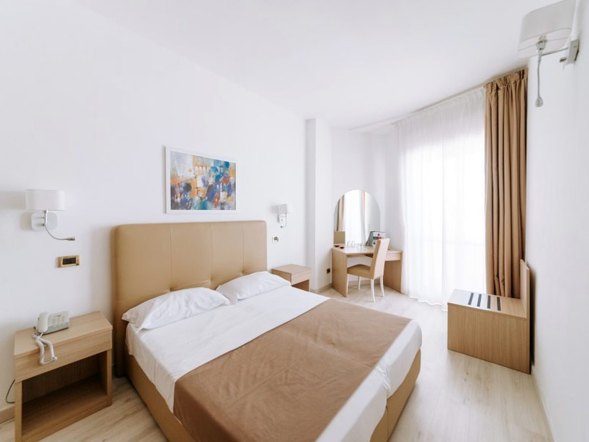 A hotel room with a double bed and padded headboard, a wooden desk and mirror, and long curtains at a balcony door.
