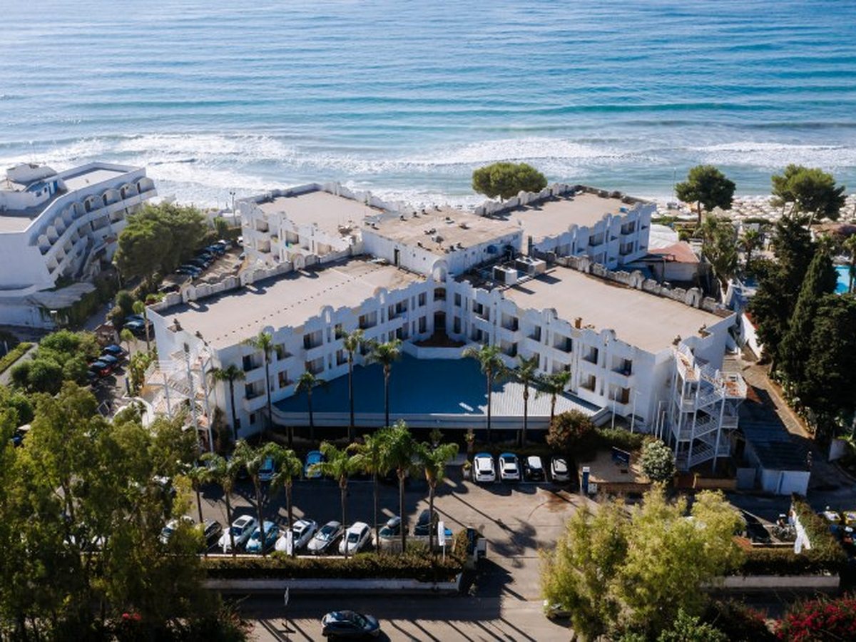The resort's white buildings from the air, with palms and the blue-roofed entrance canopy in the courtyard, a car park in front and the sea and beach behind.