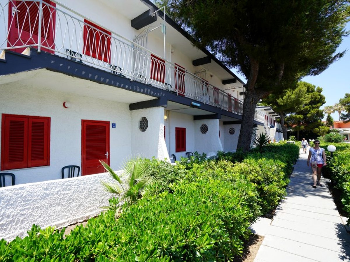 A two-storey white room block with red shutters and balconies beside a paved walkway edged with clipped hedges and pine trees.