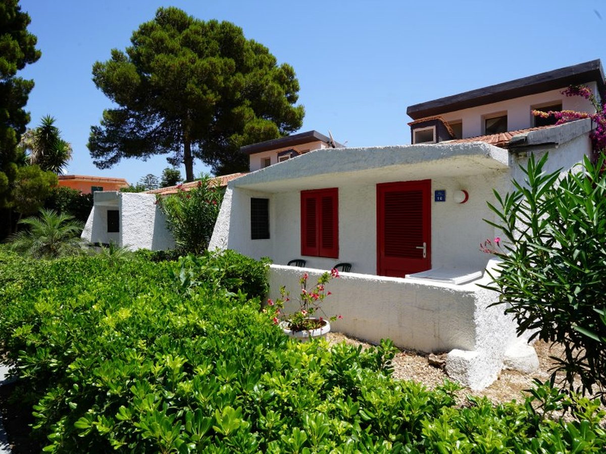 A single-storey white building with red shutters and a small walled terrace, surrounded by clipped hedges, oleander and pines under a blue sky.