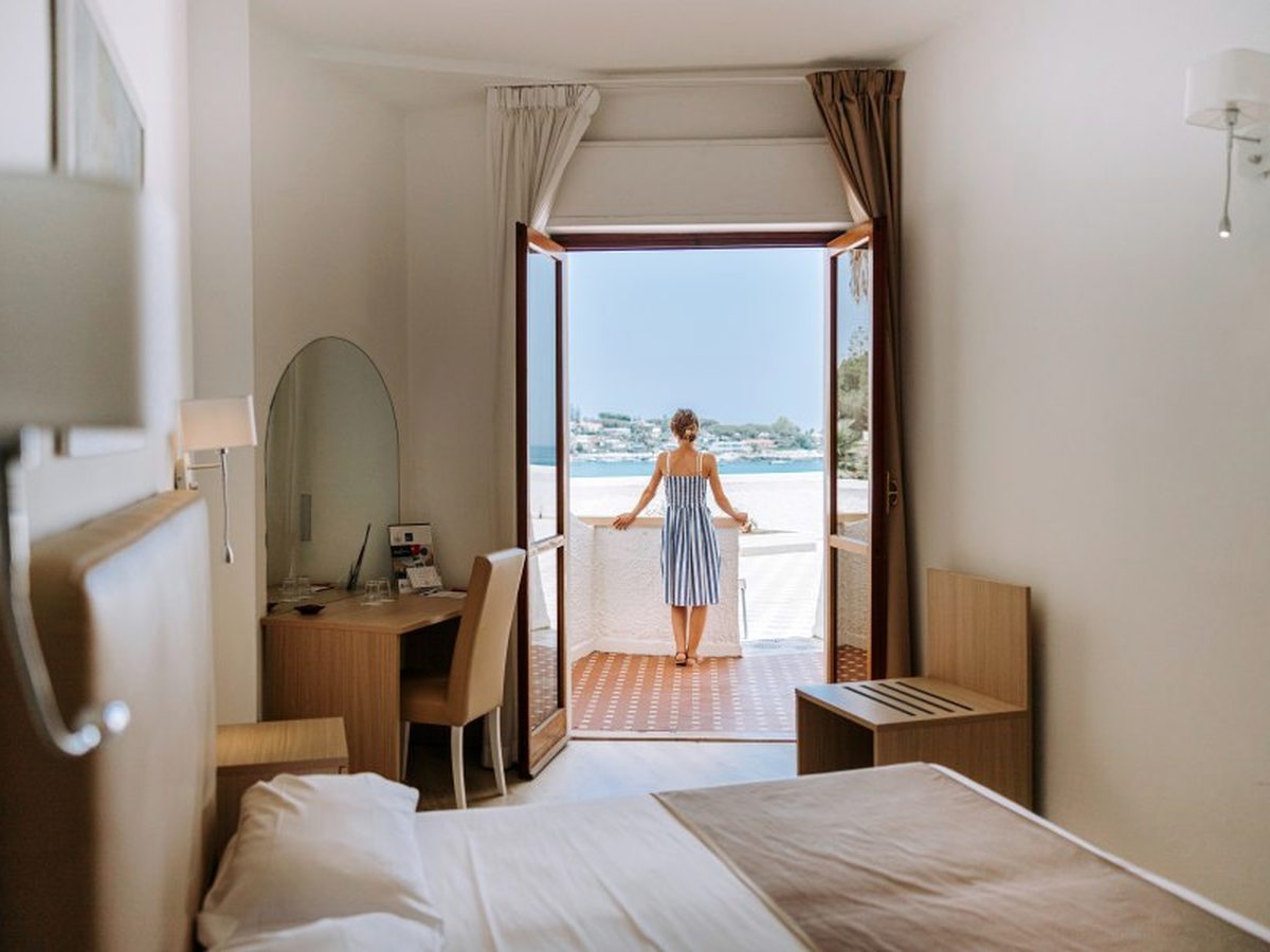 A room with its balcony doors open onto a terrace above the beach, a desk and mirror to one side and the bay beyond.