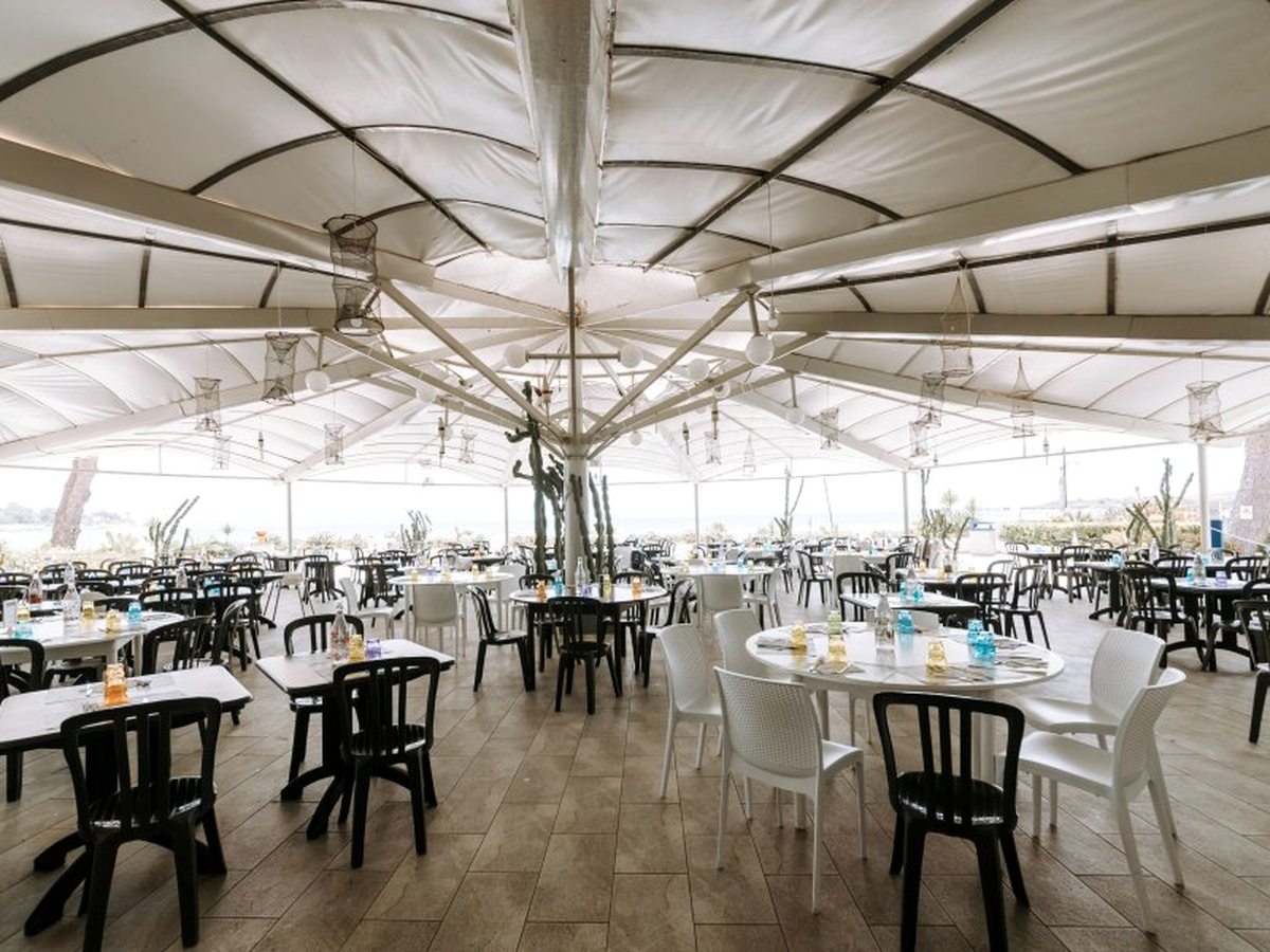 An open-air dining area under a white marquee roof hung with wicker lanterns, set with white and black tables and chairs, with the sea beyond.