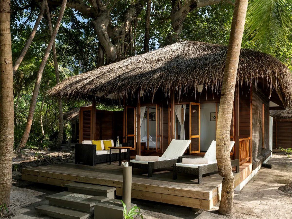 A thatched-roof villa among palm trees with a wide timber deck in front of it, two sun loungers and an outdoor sofa, and sand beyond.