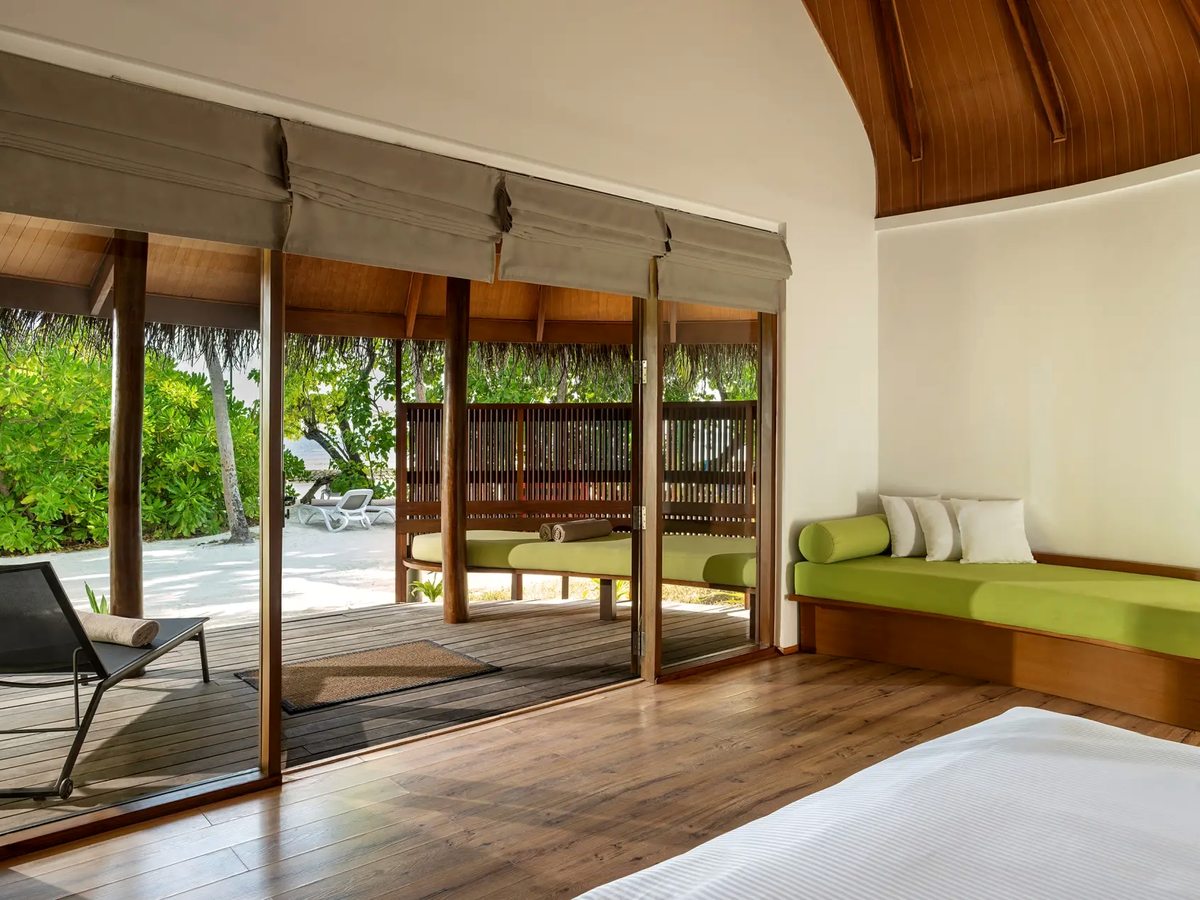 The inside of a villa with a curved wooden ceiling, a green built-in daybed along one wall and sliding doors open to a shaded deck and the beach.