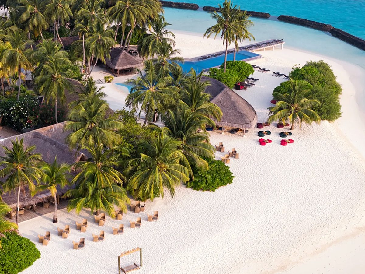 An aerial view of a wide white sand beach with rows of wicker chairs and day beds under coconut palms, thatched pavilions behind them and a pool set back among the trees.