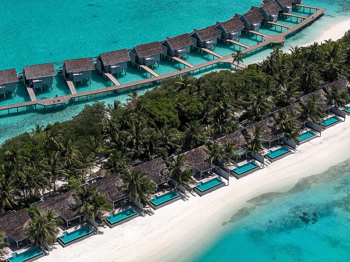 An aerial view of a stretch of the island: thatched beach villas with small private pools set behind the white sand, and a jetty of thatched overwater villas standing in the turquoise lagoon behind the palms.
