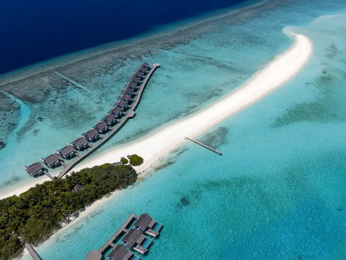 An aerial view of a long white sand spit curving off the tip of a palm-covered island into a pale turquoise lagoon, with a row of thatched overwater villas on a jetty to one side and darker reef beyond.