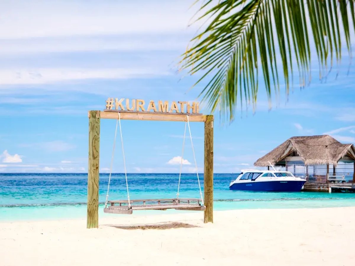A wooden swing standing on white sand at the edge of a shallow turquoise lagoon, with a thatched jetty and a moored boat behind it.