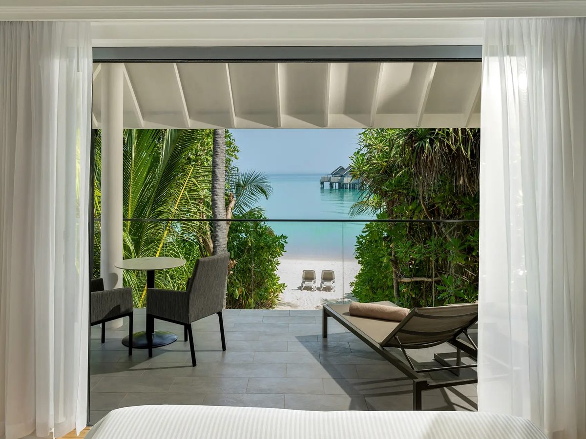 A bedroom with white curtains drawn back from open doors onto a covered terrace with sun loungers, palm trees and the turquoise lagoon beyond.