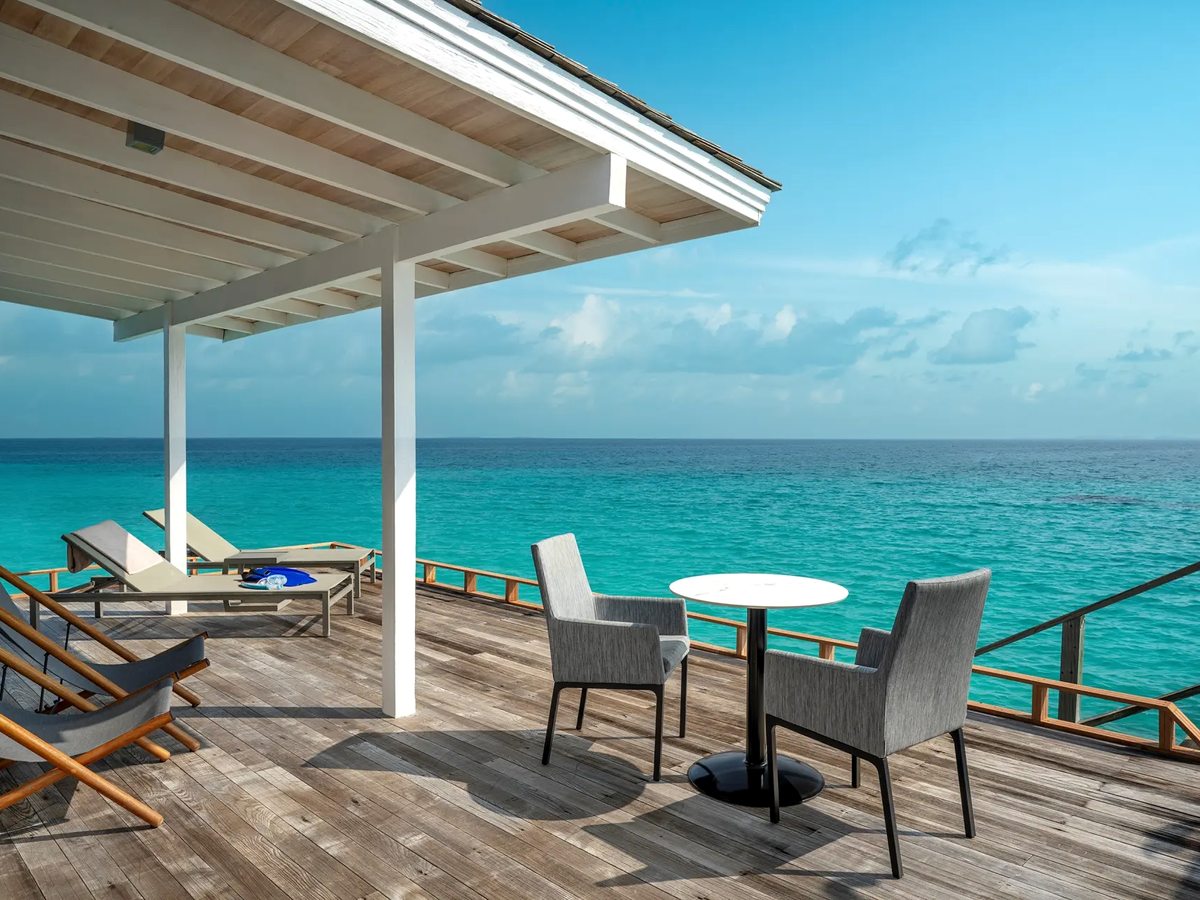 A timber deck built over clear turquoise water, with two sun loungers under a white pergola and a small table and armchairs at the rail.