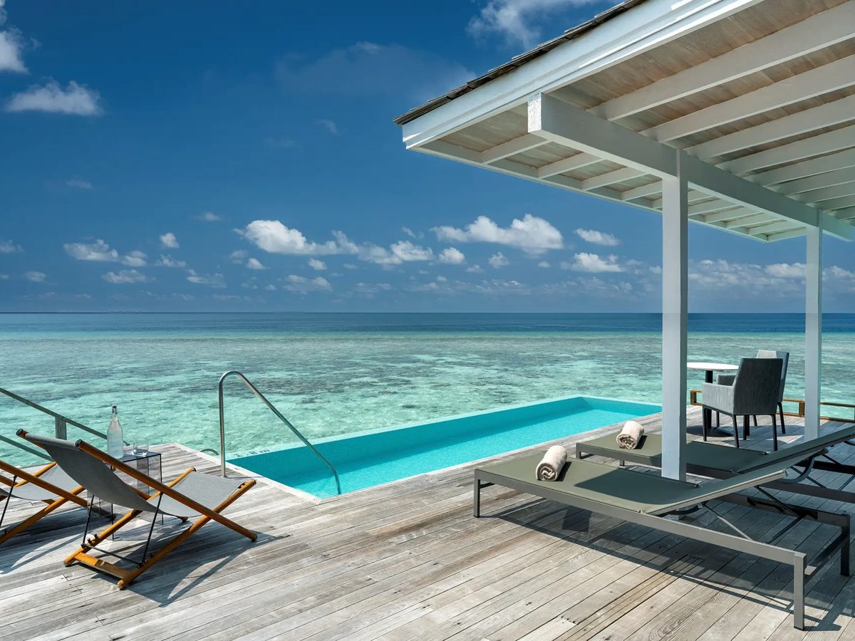A small infinity pool on a timber deck over the lagoon, with sun loungers and chairs beside it and the reef and open sea beyond.