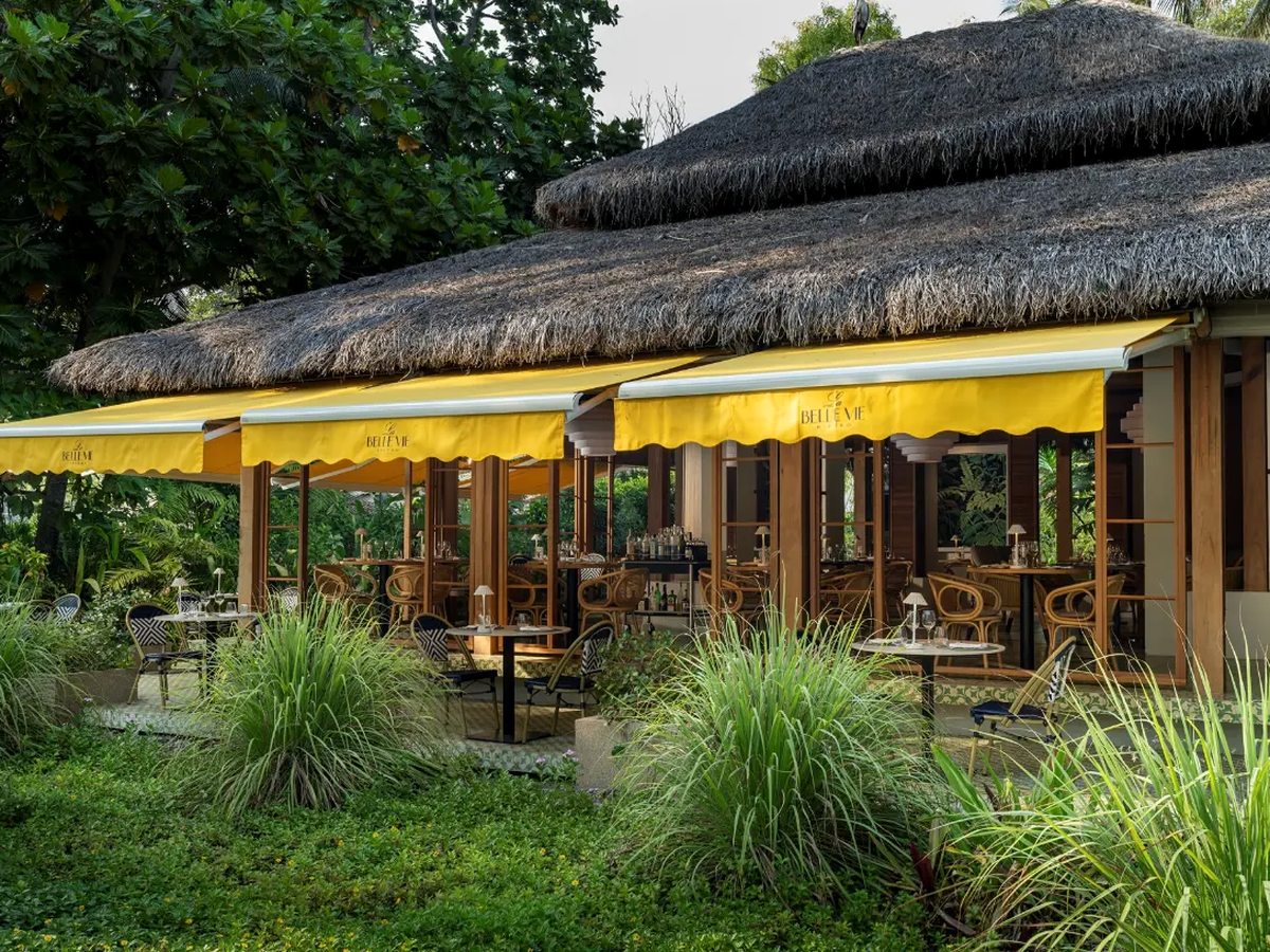 A long open-sided restaurant under a thatched roof with yellow awnings, set behind tropical planting and ornamental grasses.