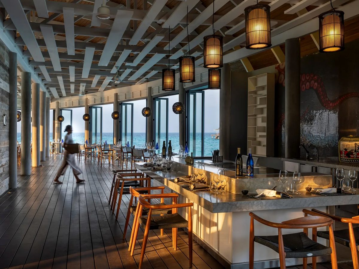A long restaurant interior with woven pendant lamps, wooden chairs and a bar counter, its full-height windows open to the sea.
