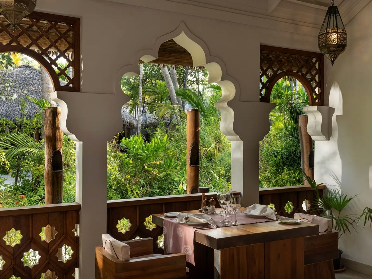 A dining table laid with glassware and pink linen inside a room with carved arched openings and lattice screens, looking into dense green garden.