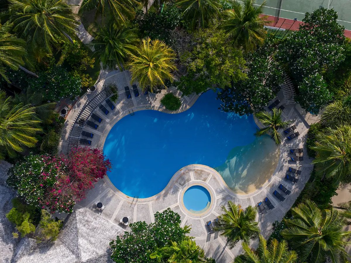 An aerial view of a curving freeform pool with a small round pool beside it, ringed by sun loungers, palms and flowering shrubs, with a tennis court at one corner.