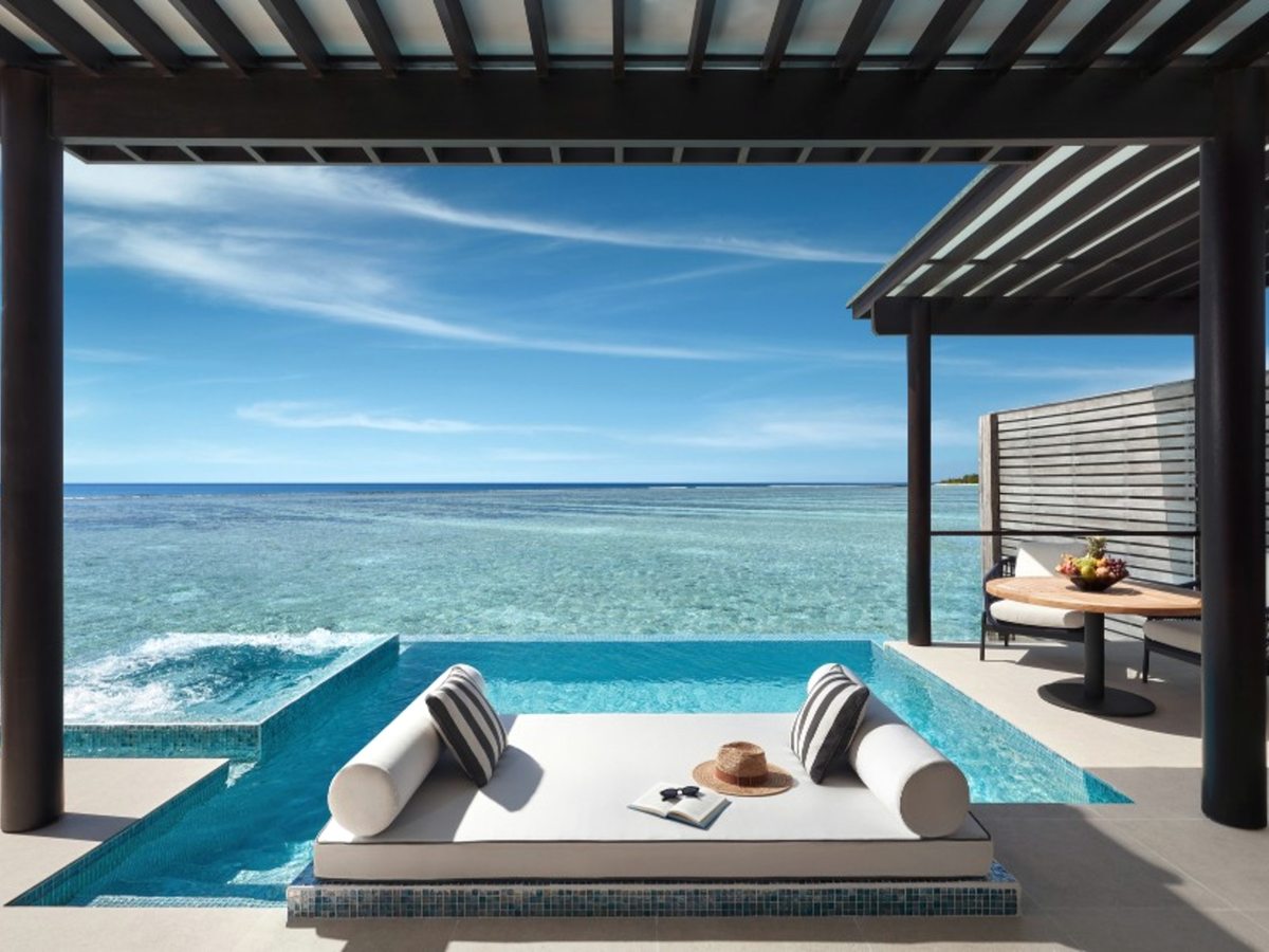 A private infinity pool on an overwater villa deck with a cushioned double daybed at its edge, under a dark timber pergola, above a shallow turquoise lagoon.