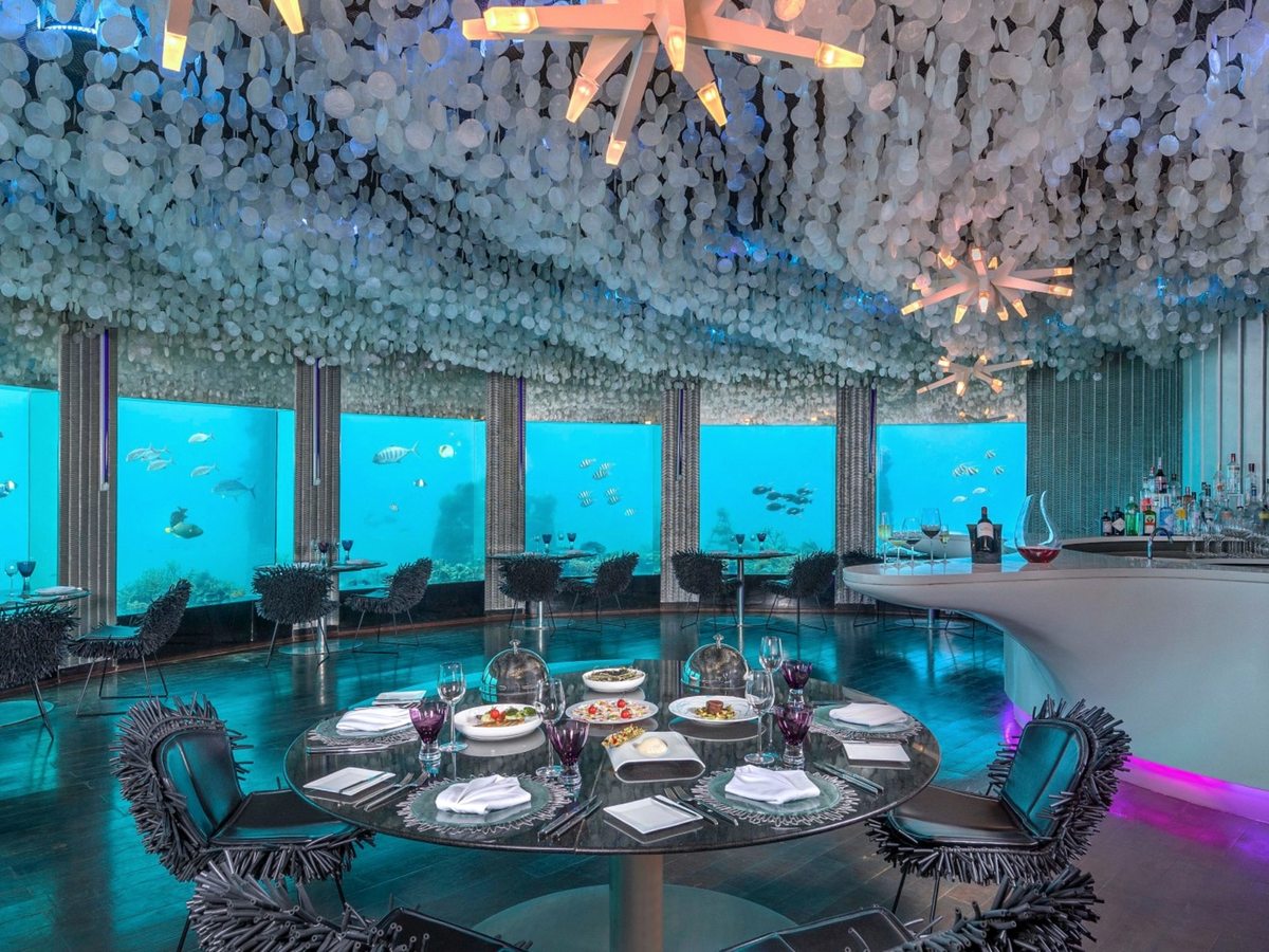 An underwater dining room with curved windows onto a coral reef and shoals of fish, a laid round table in the centre, spiked designer chairs and a shell-strung ceiling.