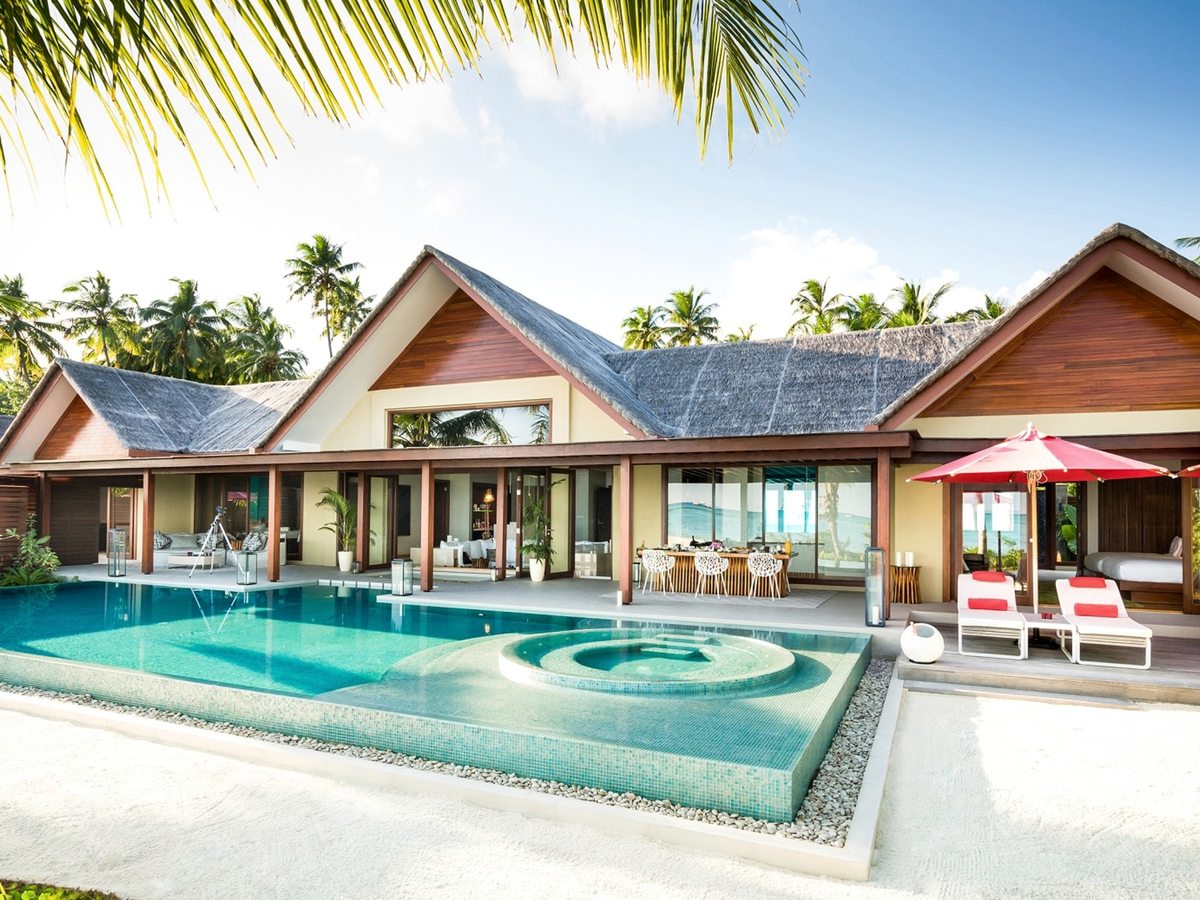 A large thatched villa with open glass frontage behind a full-sized rectangular pool with a circular jacuzzi set into one end, sun loungers under a red parasol and white sand around it.
