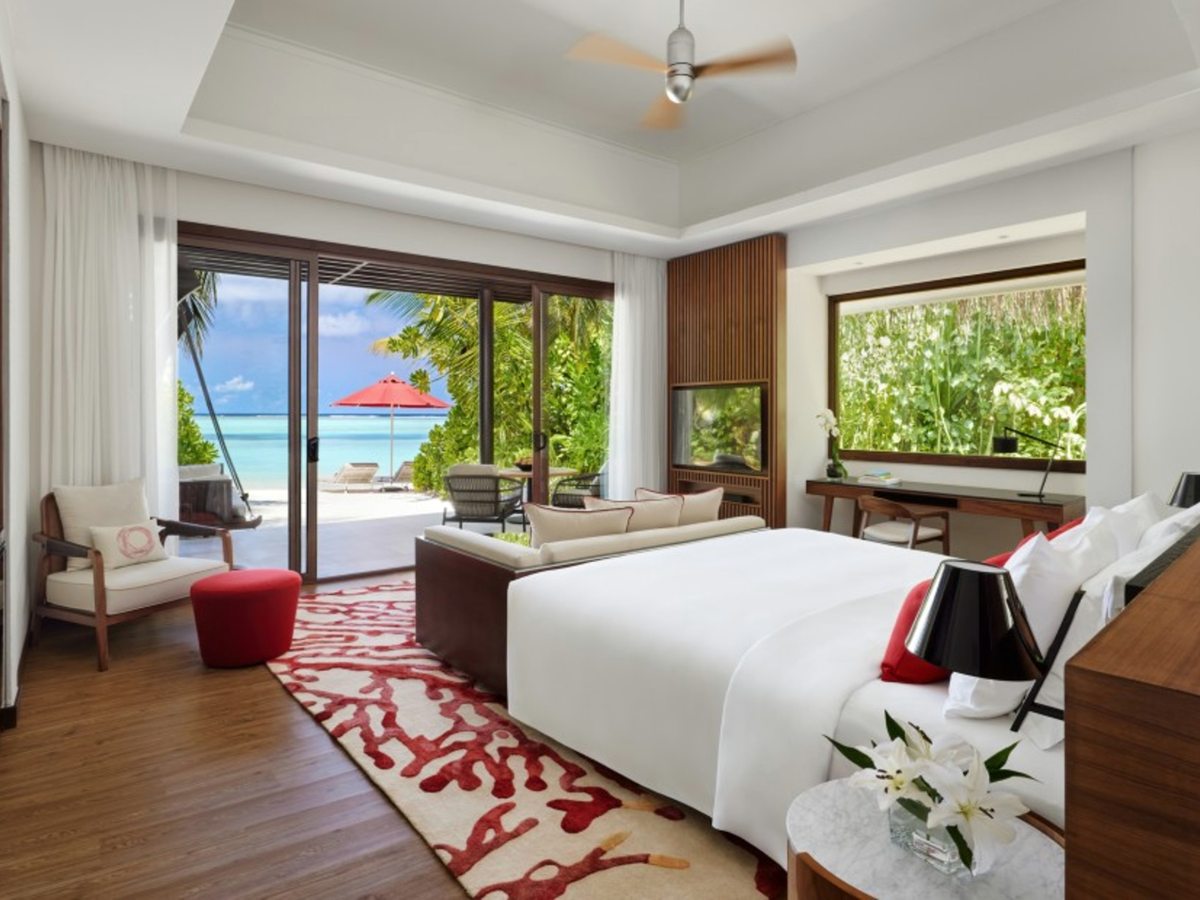 A villa bedroom with a king bed, red coral-patterned rug, armchair and desk, with sliding doors open onto white sand, a red parasol and the turquoise sea.
